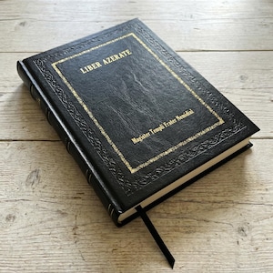 Könnte beinhalten: Schwarzes, ledergebundenes Notizbuch mit goldfarbenen Buchstaben und Verzierungen. Auf dem Einband steht "LIBER AZERATE" und "Magister Templi Frater Nemidial". Ein schwarzes Leseband ist zu sehen.