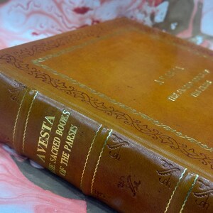 May include: Close-up of a vintage leather-bound book titled "Avesta: The Sacred Books of the Parsis." The cover is a warm brown with gold lettering and decorative accents. The book's spine features vertical gold lines.