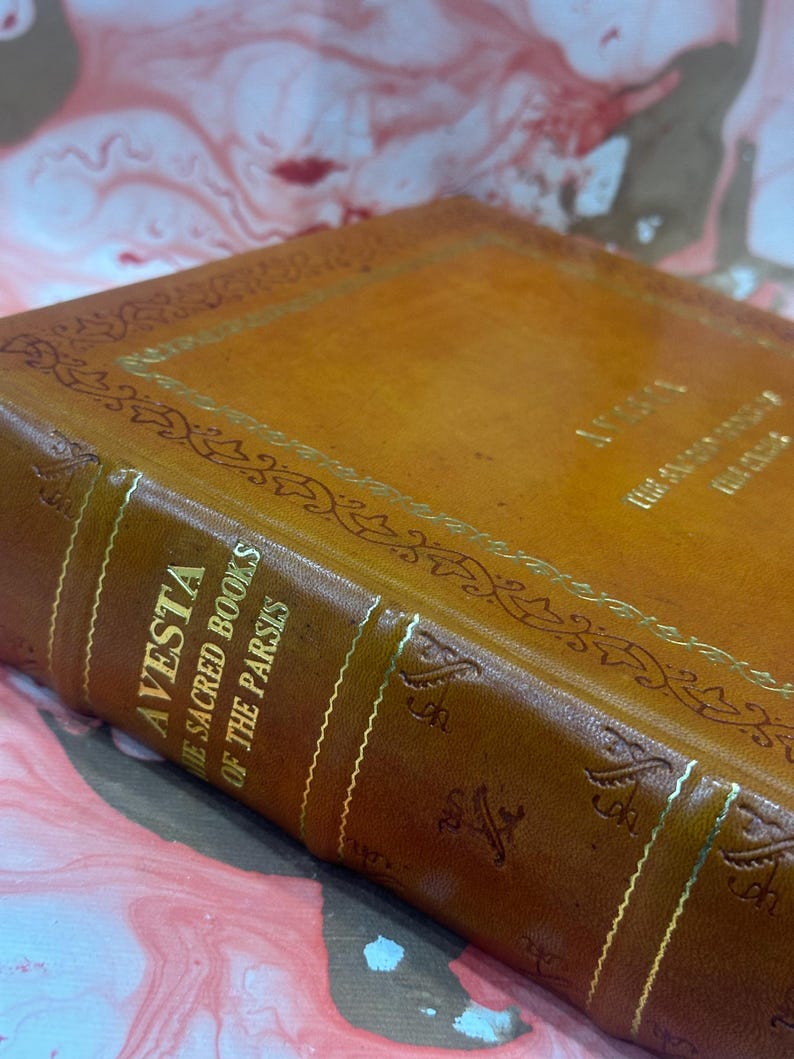 May include: Close-up of a vintage leather-bound book titled "Avesta: The Sacred Books of the Parsis." The cover is a warm brown with gold lettering and decorative accents. The book's spine features vertical gold lines.