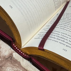 May include: An open book with text visible on the pages, displaying page numbers 152 and 153. The book features a burgundy cover and gold-toned edges. A burgundy ribbon bookmark is placed between the pages.