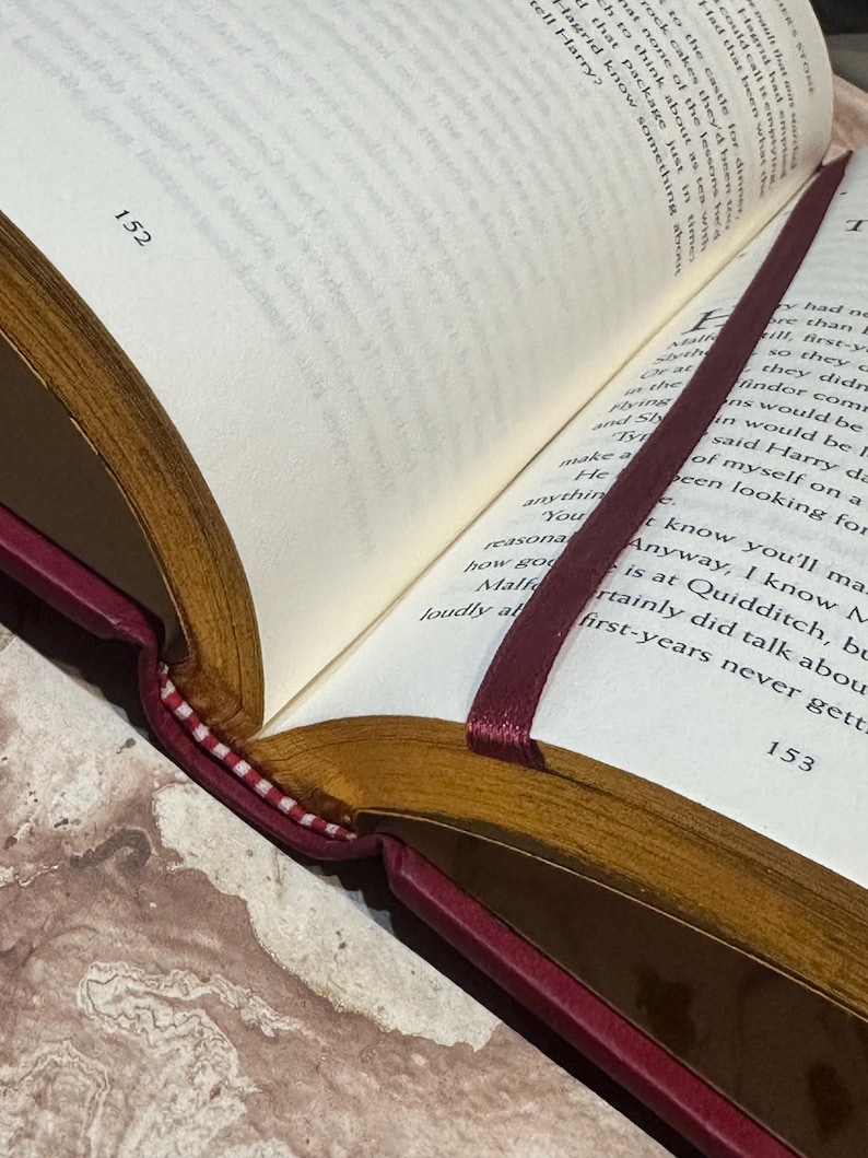 May include: An open book with text visible on the pages, displaying page numbers 152 and 153. The book features a burgundy cover and gold-toned edges. A burgundy ribbon bookmark is placed between the pages.