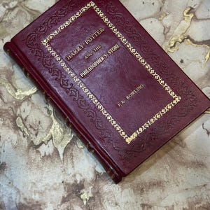 Puede incluir: Un libro de tapa dura burdeos titulado "Harry Potter and the Philosopher's Stone" con letras doradas y un borde decorativo. El nombre del autor, J.K. Rowling, también está en oro. El libro está cerrado.