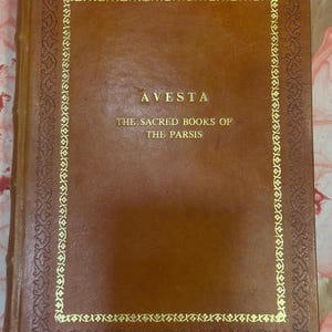 May include: A brown leather-bound book titled "AVESTA" with gold lettering and a decorative gold border. The text below reads "THE SACRED BOOKS OF THE PARSIS". The book's cover is a solid, rich brown.