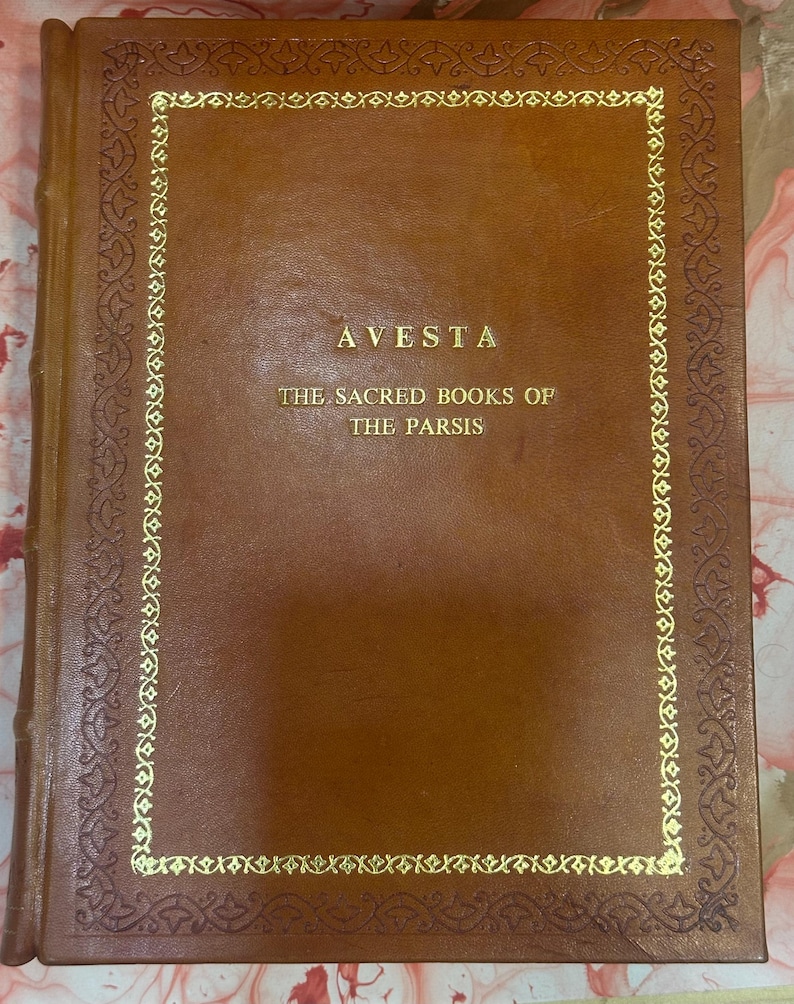 May include: A brown leather-bound book titled "AVESTA" with gold lettering and a decorative gold border. The text below reads "THE SACRED BOOKS OF THE PARSIS". The book's cover is a solid, rich brown.