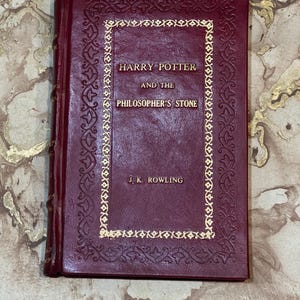 May include: A burgundy hardback book titled "Harry Potter and the Philosopher's Stone" by J.K. Rowling. The cover has gold lettering and a decorative gold border. The book is closed and appears to be in good condition.