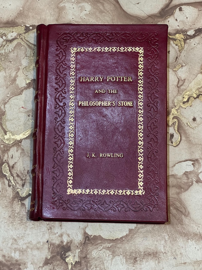 May include: A burgundy hardback book titled "Harry Potter and the Philosopher's Stone" by J.K. Rowling. The cover has gold lettering and a decorative gold border. The book is closed and appears to be in good condition.