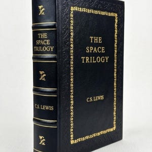 May include: A black hardcover book titled "The Space Trilogy" by C.S. Lewis, with gold lettering and decorative borders. The spine also features gold lettering and embellishments. A classic trilogy.