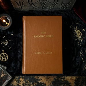 May include: A brown hardcover book titled "The Satanic Bible" by Anton S. LaVey, with gold lettering. The book is on a black surface, surrounded by a lit candle, tarot cards, and a pentagram pendant.