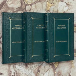 May include: Three hardback books with green covers and gold lettering. The titles are "Howl's Moving Castle", "Castle in the Air", and "House of Many Ways" by Diana Wynne Jones. The books have decorative gold borders.