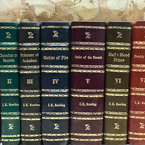 May include: A set of seven hardback books, featuring titles such as "Philosopher's Stone" and "Deathly Hallows." The spines are in shades of red, green, and navy, with gold lettering and decorative details. Each spine displays the author's name, J.K. Rowling.