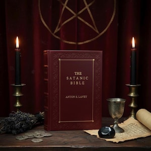 May include: A burgundy-colored book titled "The Satanic Bible" by Anton S. LaVey, displayed on a wooden surface. Two black candles in brass holders flank the book. A silver goblet, coins, and a scroll are also present.