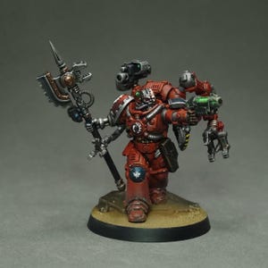 Puede incluir: Figura en miniatura de un guerrero con armadura roja, equipado con un hacha mecánica y una pistola láser verde. La figura tiene un casco con forma de calavera y está sobre una base redonda.