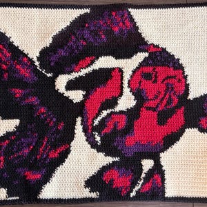 May include: Handmade crochet blanket featuring a colorful abstract design. The blanket showcases a bold pattern with black, red, purple, and cream-colored yarn. The design appears to be a stylized figure or shape.