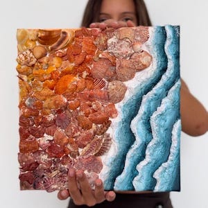 May include: Square artwork depicting a beach scene made of seashells. The left side is composed of orange and brown shells, transitioning to a blue and white wave design on the right. The artwork is held up by a person.