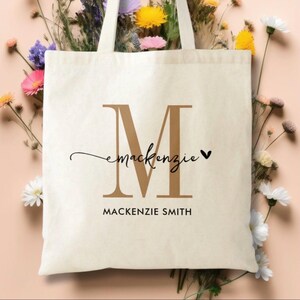 Pu&ograve; includere: Borsa tote color crema con una grande lettera "M" marrone e il nome "Mackenzie" in corsivo. Sotto, il nome "Mackenzie Smith" &egrave; stampato in un semplice font nero. La borsa &egrave; circondata da fiori colorati.