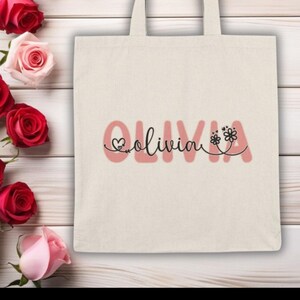 Pu&ograve; includere: Una borsa tote color crema con il nome "Olivia" in corsivo, decorata con un cuore e motivi floreali. La borsa &egrave; progettata per trasportare vari oggetti. Rose rosse e rosa sullo sfondo.