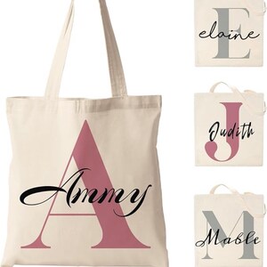 Pu&ograve; includere: Borse tote color crema con nomi personalizzati. Una borsa presenta il nome "Ammy" con una grande lettera "A" rosa. Altre borse mostrano i nomi "Elaine", "Judith" e "Mable" in caratteri eleganti.
