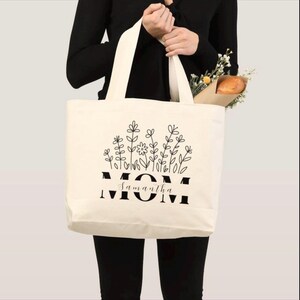 Pu&ograve; includere: Borsa tote color crema con design floreale nero e la parola "MOM" in grassetto, con "Samantha" sotto. La borsa ha manici lunghi ed &egrave; piena di fiori e una baguette.