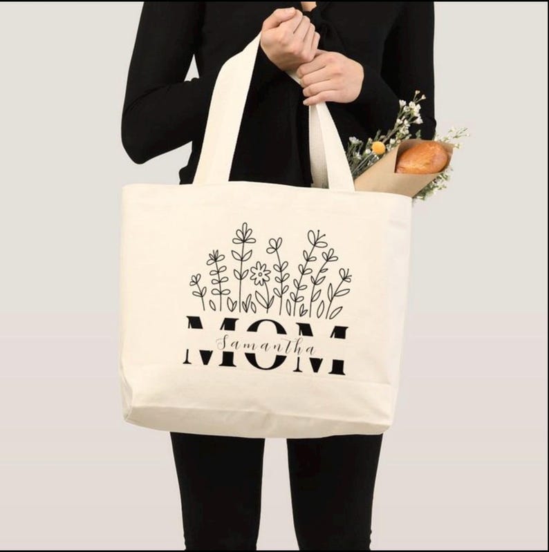 Pu&ograve; includere: Borsa tote color crema con design floreale nero e la parola "MOM" in grassetto, con "Samantha" sotto. La borsa ha manici lunghi ed &egrave; piena di fiori e una baguette.