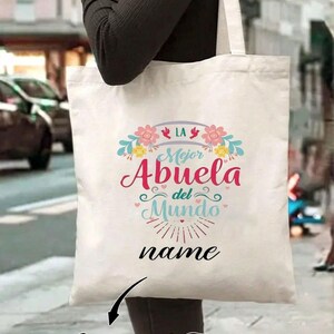 Pu&ograve; includere: Una borsa tote bianca con la scritta "LA Mejor Abuela del Mundo" in rosa e blu, con decorazioni floreali. La borsa presenta anche la parola "name" e "Customized Text" in nero.