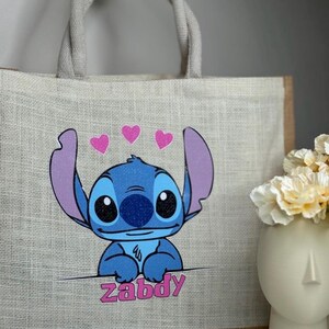Pu&ograve; includere: Una borsa tote in iuta beige con un'immagine a cartoni animati di Stitch, un personaggio alieno blu, e il nome "Zabdy" in rosa. La borsa ha un bordo marrone, manici in corda grigia e tre cuori rosa.
