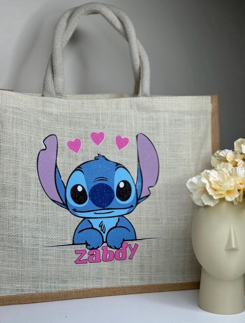 Pu&ograve; includere: Una borsa tote in iuta beige con un'immagine a cartoni animati di Stitch, un personaggio alieno blu, e il nome "Zabdy" in rosa. La borsa ha un bordo marrone, manici in corda grigia e tre cuori rosa.