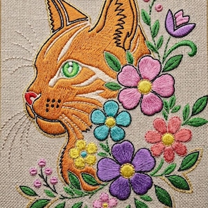 May include: An embroidery piece on beige fabric featuring an orange cat head with bright green eyes, surrounded by a colorful arrangement of embroidered pink, purple, blue, and yellow flowers with green leaves and small pink buds.