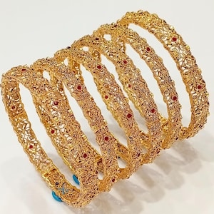 Puede incluir: Un conjunto de seis brazaletes dorados con un intrincado patrón de filigrana metálica. Cada pulsera circular está acentuada con pequeñas piedras rojas espaciadas uniformemente, incrustadas en el diseño calado y texturizado del metal.