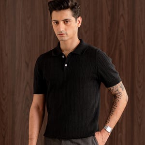 May include: Black short-sleeved polo shirt with a cable knit texture. The shirt features a classic collar and two buttons. The model is wearing grey trousers and a silver wristwatch.
