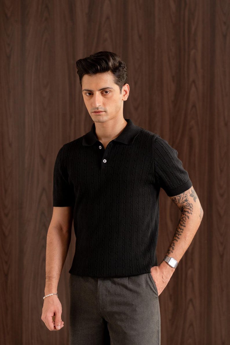 May include: Black short-sleeved polo shirt with a cable knit texture. The shirt features a classic collar and two buttons. The model is wearing grey trousers and a silver wristwatch.