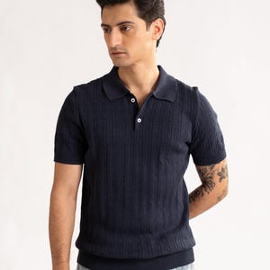 May include: A navy blue polo shirt with a cable knit pattern. The short-sleeved top features a collared neckline and two white buttons. The shirt is worn with light blue jeans. The style is casual and smart.