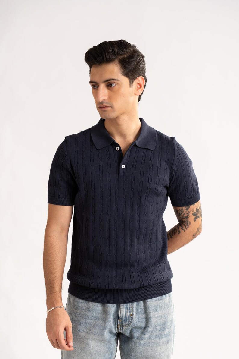 May include: A navy blue polo shirt with a cable knit pattern. The short-sleeved top features a collared neckline and two white buttons. The shirt is worn with light blue jeans. The style is casual and smart.