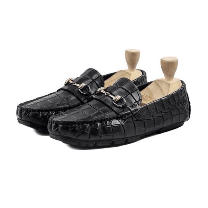 May include: A pair of black leather loafers with a crocodile pattern. Each shoe features a gold-colored metal buckle and is displayed with a wooden shoe tree. The shoes are designed for comfort and style.