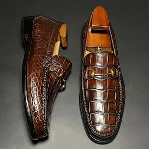 May include: A pair of brown leather loafers with a crocodile skin texture. The shoes have a gold-colored metal buckle and a wooden shoe tree inside. The loafers are displayed on a dark surface.