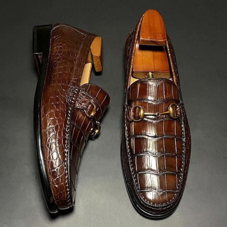 May include: A pair of brown leather loafers with a crocodile skin texture. The shoes have a gold-colored metal buckle and a wooden shoe tree inside. The loafers are displayed on a dark surface.