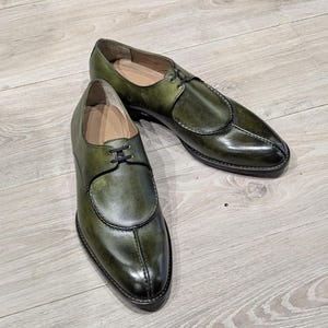 Peut inclure: Une paire de chaussures habillées en cuir vert olive avec une finition brillante. Les chaussures présentent un design unique avec un bout arrondi et des lacets noirs. Les chaussures sont présentées sur une surface en bois clair.