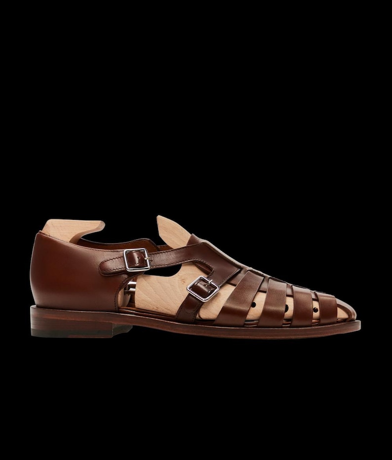 May include: Brown leather sandal with multiple straps and silver buckles. The shoe has a low heel and a wooden shoe form inside. The sandal is designed with a cage-like structure.