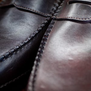 May include: Close-up of two brown leather penny loafers. The shoes are stitched with a visible seam and have a glossy finish. The image highlights the texture and craftsmanship of the footwear, showcasing the classic design.