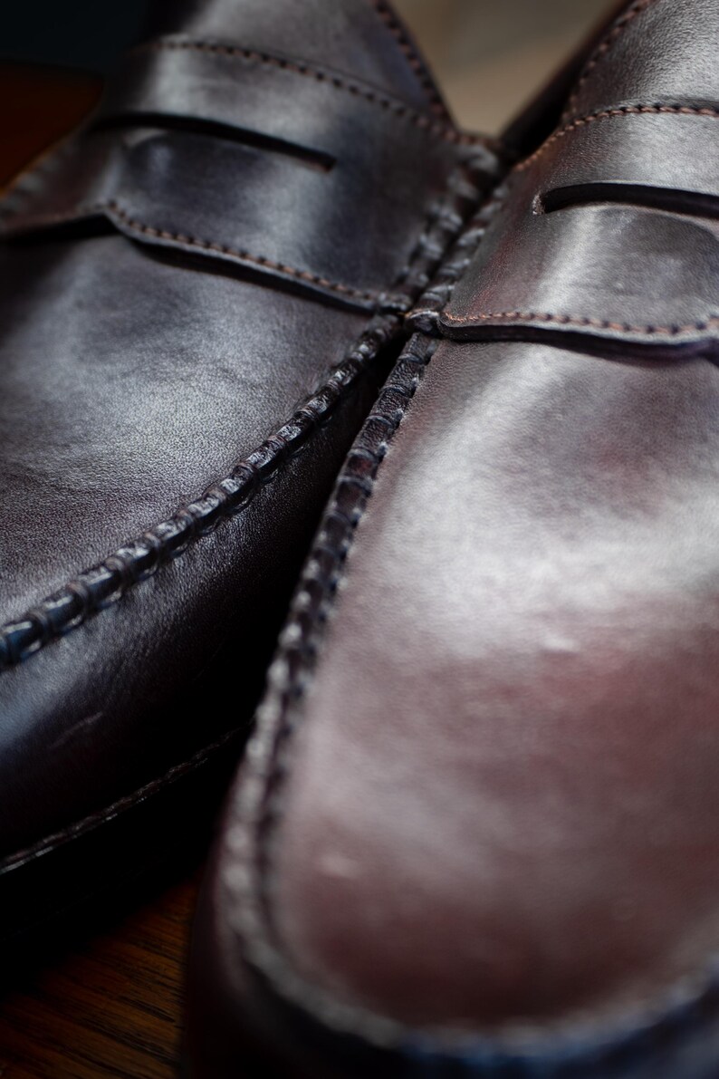 May include: Close-up of two brown leather penny loafers. The shoes are stitched with a visible seam and have a glossy finish. The image highlights the texture and craftsmanship of the footwear, showcasing the classic design.