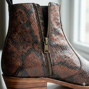 May include: A close-up of a brown and black ankle boot with a snakeskin pattern. The boot has a side zipper with a brass-colored pull. The heel is a light brown color.