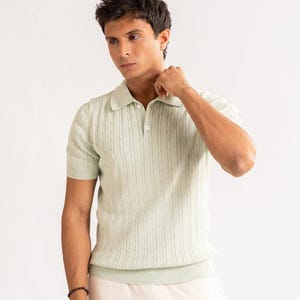 May include: A pale green polo shirt with a cable knit pattern. The shirt features short sleeves and a collared neckline with two buttons. The model is wearing a black bracelet and cream-colored trousers. The background is white.
