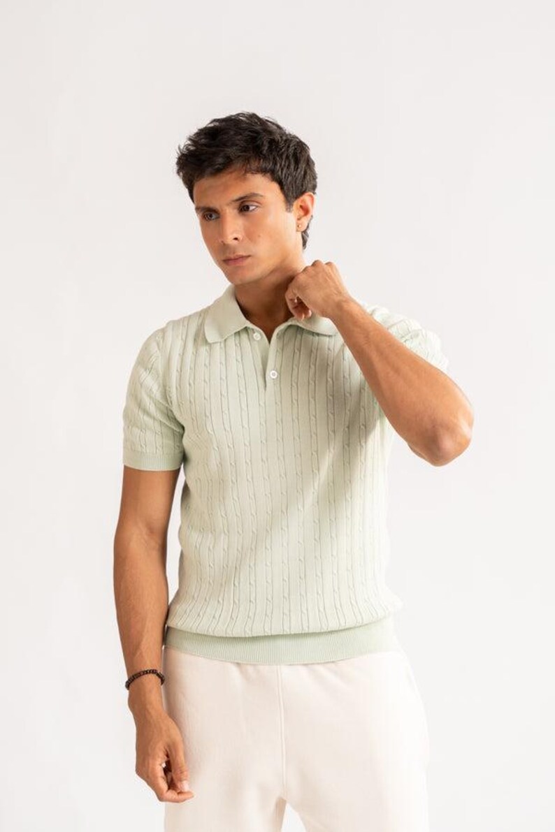 May include: A pale green polo shirt with a cable knit pattern. The shirt features short sleeves and a collared neckline with two buttons. The model is wearing a black bracelet and cream-colored trousers. The background is white.