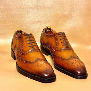 Puede incluir: Un par de zapatos brogue de cuero marr&oacute;n con cordones marrones oscuros. Los zapatos tienen un dise&ntilde;o cl&aacute;sico de punta de ala con perforaciones decorativas y una suela marr&oacute;n oscuro. El cuero tiene un acabado pulido y brillante.