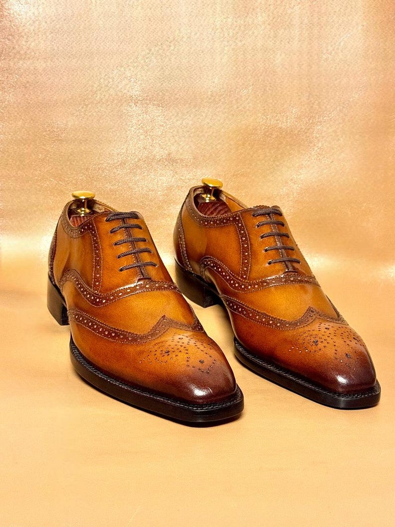 Puede incluir: Un par de zapatos brogue de cuero marr&oacute;n con cordones marrones oscuros. Los zapatos tienen un dise&ntilde;o cl&aacute;sico de punta de ala con perforaciones decorativas y una suela marr&oacute;n oscuro. El cuero tiene un acabado pulido y brillante.