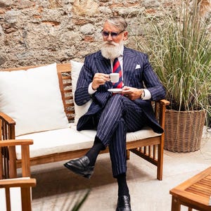 Pu&ograve; includere: Un uomo in un abito gessato blu navy con cravatta a righe rosse e blu, scarpe nere e occhiali da sole, tiene in mano una tazza e un piattino bianchi. &Egrave; seduto su una panchina di legno con un cuscino bianco, con un muro di mattoni sullo sfondo.