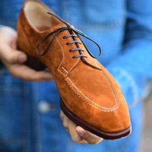 Op de afbeelding: Bruine suède herenschoenen met donkerbruine veters en stiksels. De schoenen hebben een klassiek design met een ronde neus en een leren zool. De schoenen worden in iemands handen gehouden tegen een blauwe achtergrond.