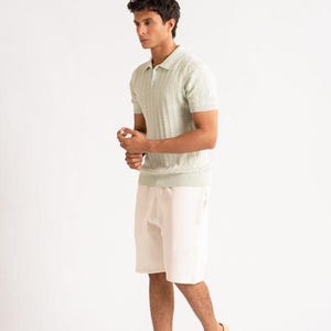 May include: A pale green knitted polo shirt with a textured design, worn with cream shorts and beige loafers. The person is standing against a white backdrop. The outfit is a casual summer look.