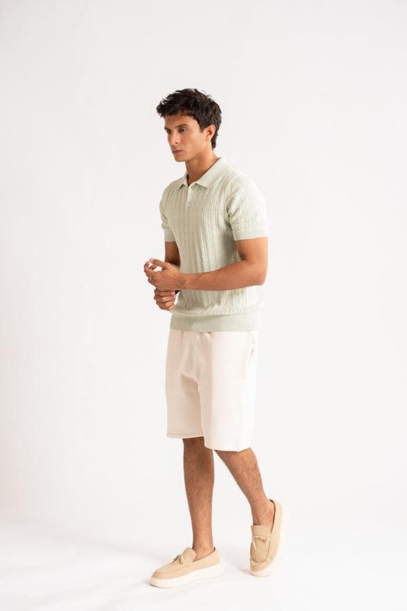May include: A pale green knitted polo shirt with a textured design, worn with cream shorts and beige loafers. The person is standing against a white backdrop. The outfit is a casual summer look.