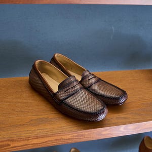 May include: A pair of dark brown loafers featuring a textured, woven-style grid pattern across the upper surface. The shoes have a classic slip-on design with a rounded toe, a horizontal strap across the vamp, and a smooth, light tan interior lining.