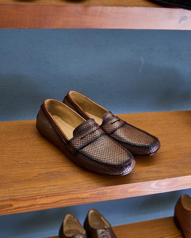 May include: A pair of dark brown loafers featuring a textured, woven-style grid pattern across the upper surface. The shoes have a classic slip-on design with a rounded toe, a horizontal strap across the vamp, and a smooth, light tan interior lining.